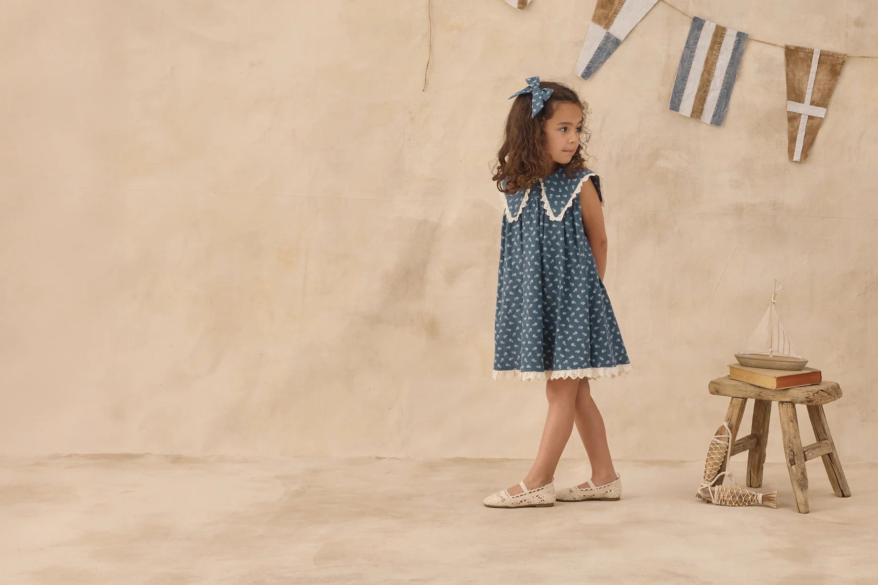 Young girl in a polka dot dress standing in front of a wooden stool with a toy boat on it, against a beige wall.
