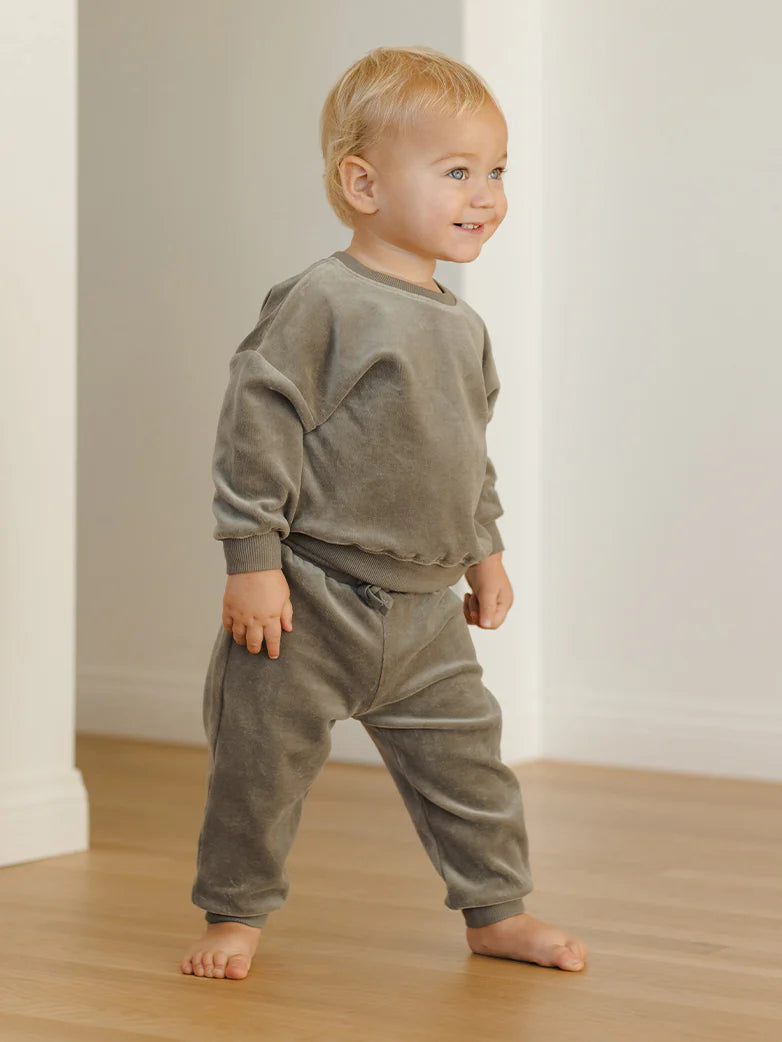 Child wearing a green outfit standing on a wooden floor with a white wall background
